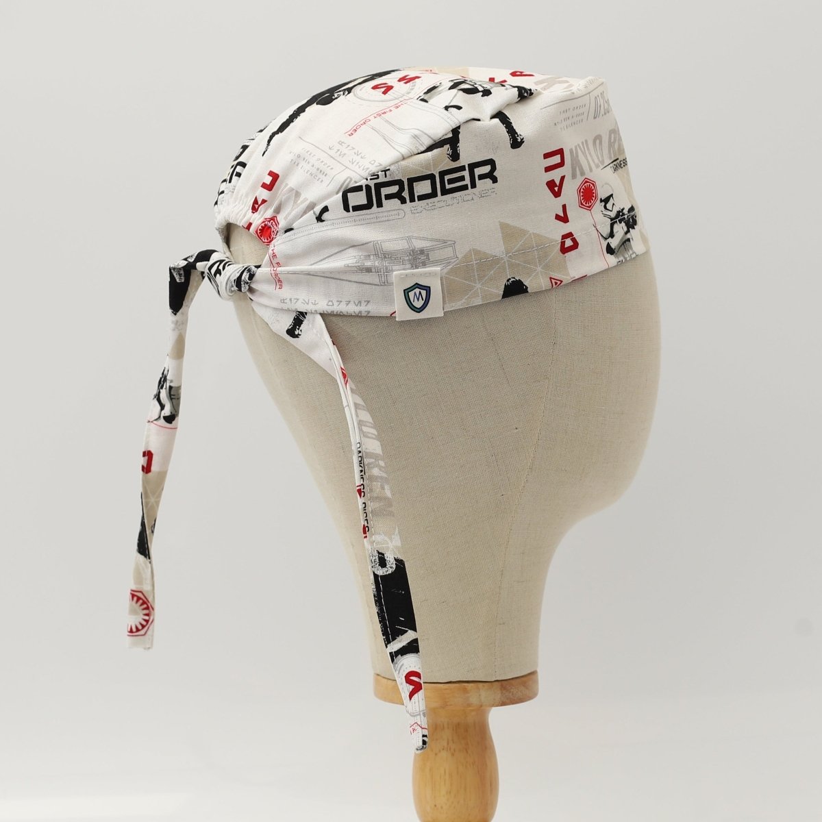 Custom white scrub cap by Medicus Caps featuring black and red geometric shapes and futuristic text pattern; side/back view on mannequin with long ties.