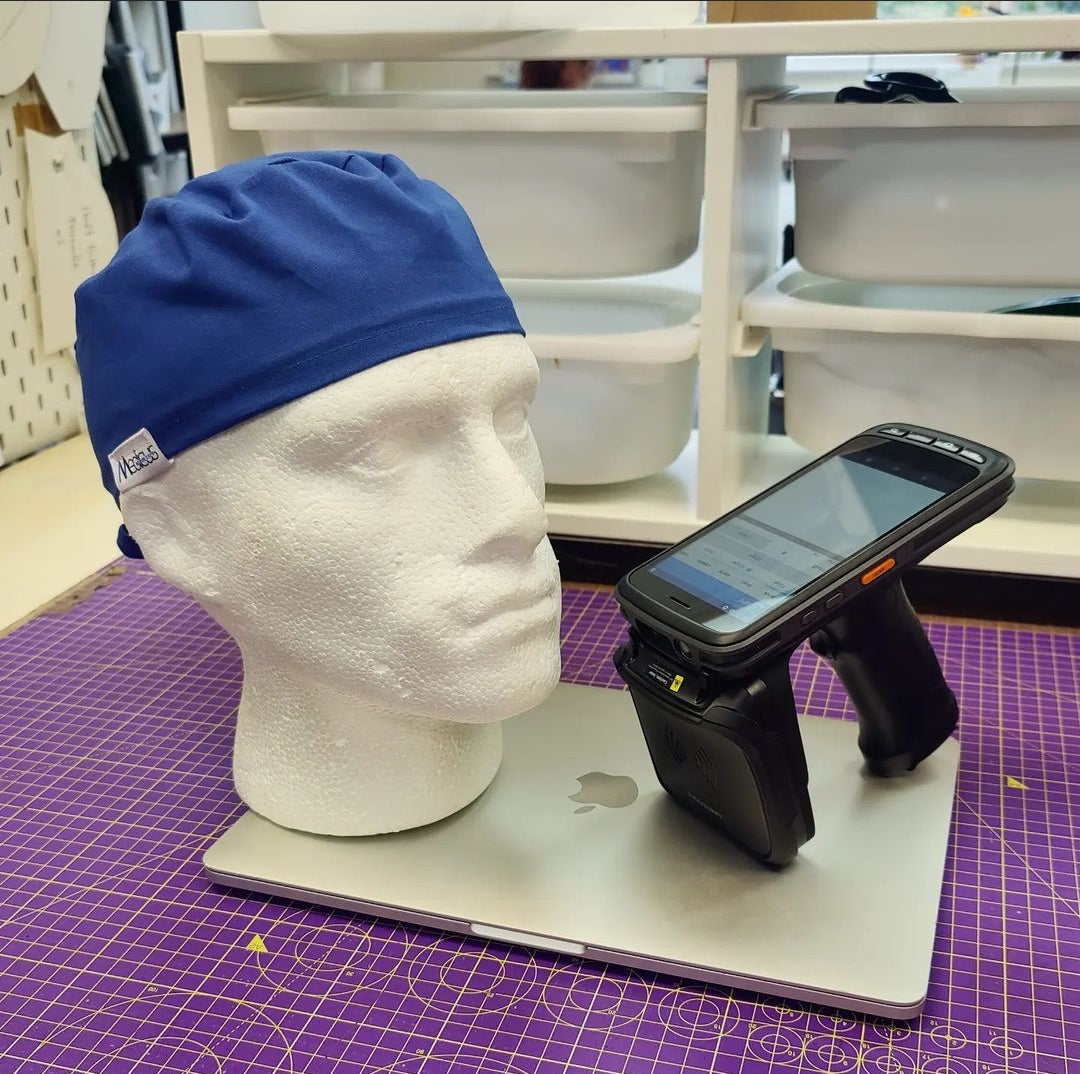A mannequin head with a blue cap sits on a laptop, beside a handheld barcode scanner resting on the laptop. The background shows shelves with stacked items.