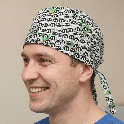 Black and white cartoon character pattern scrub cap with green accents worn by smiling healthcare worker