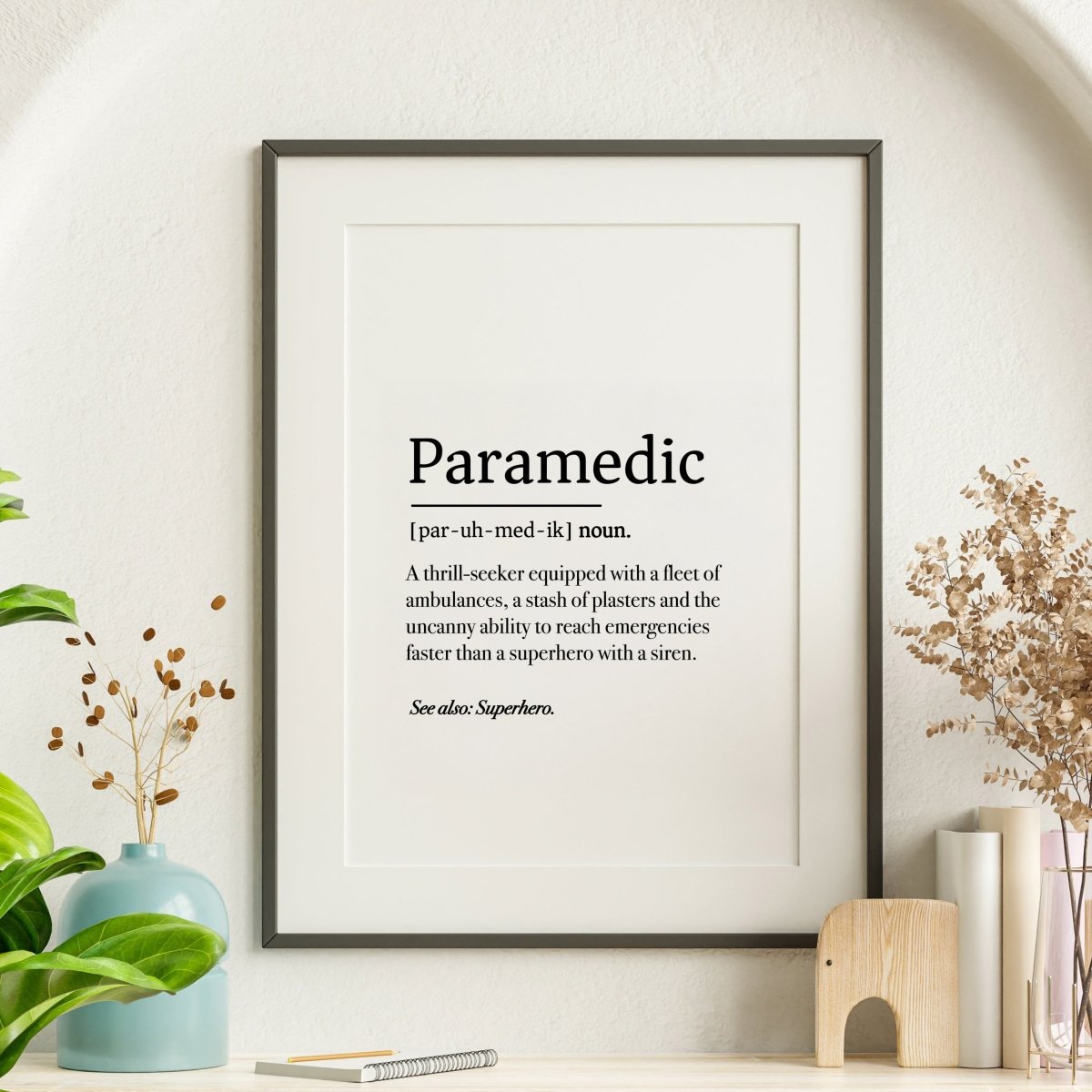 The Paramedic Definition poster by Medicus Scrub Caps humorously defines paramedics as superheroes with sirens. Displayed in a frame with plants and decor.