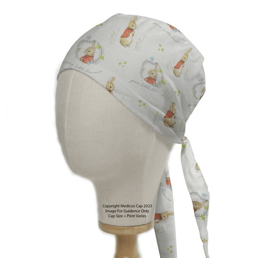 A mannequin head displays a Peter Rabbit Grey Cartoon Scrub Cap by Medicus Scrub Caps, featuring rabbits and green flowers. The cap has a back tie and printed text with copyright and size details, reminiscent of Beatrix Potters tales.