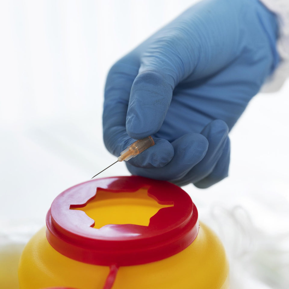 Gloved hand disposing a used syringe into a yellow clinical sharps waste bin
