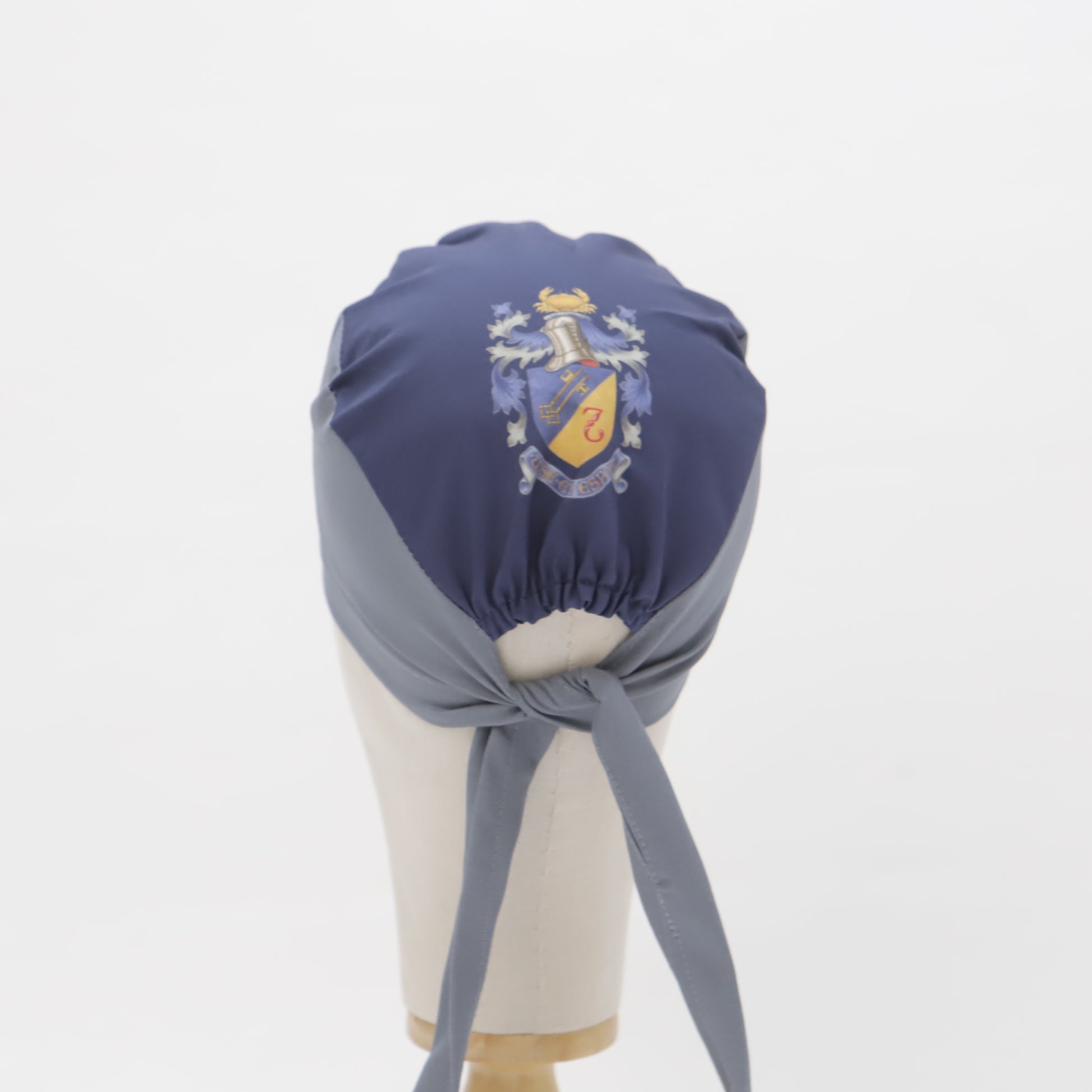 A wig head displays a Specific Custom Printed Placement Scrub Cap by Medicus Scrub Caps, featuring a detailed coat of arms on dark blue fabric.