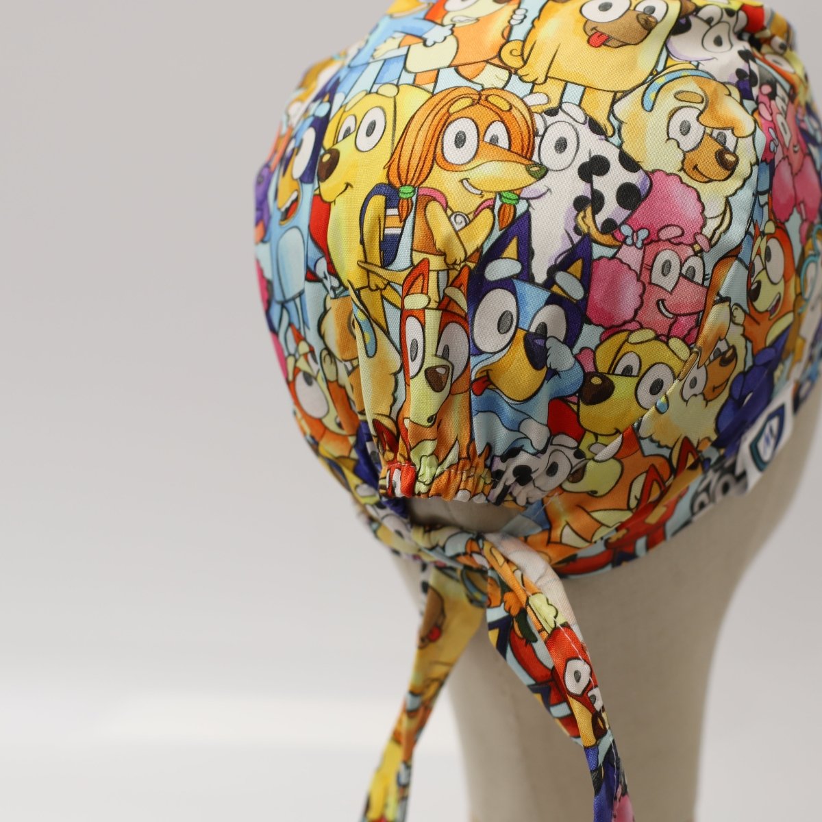 Custom scrub cap featuring colorful cartoon animals and characters print on a white background; handmade by Medicus Caps. Side/back view shows ties and bouffant style.