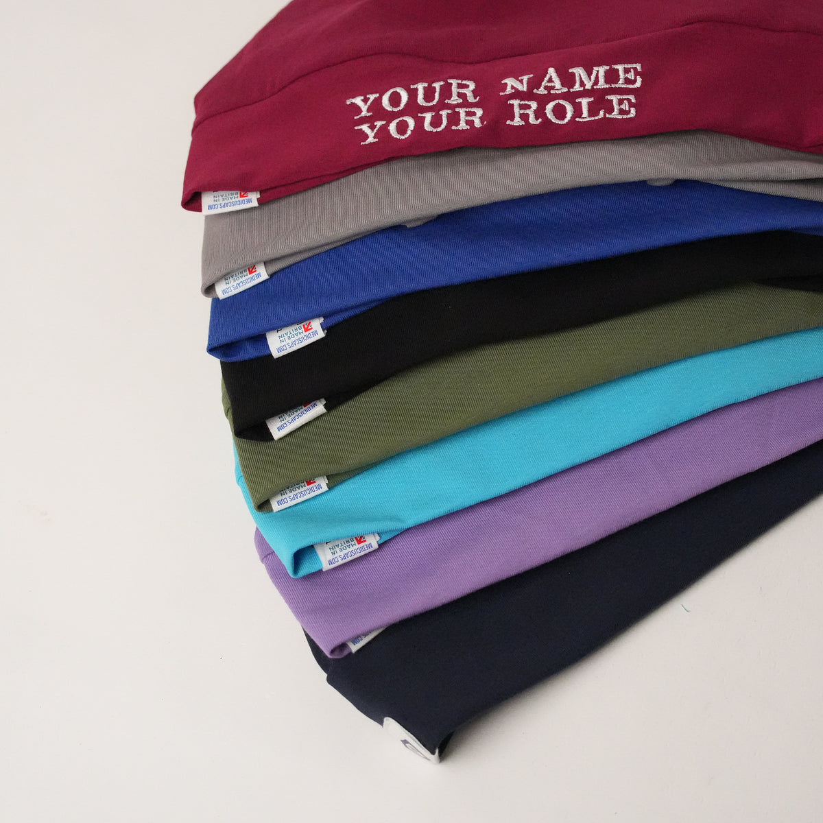 Stack of seven Contour Plain Scrub Caps in burgundy, grey, blue, black, olive, cyan and purple, personalised cap on top.