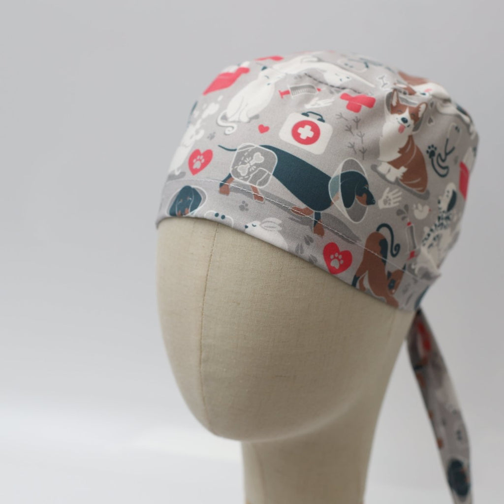 Side view of a grey custom scrub cap featuring dachshund dogs and red hearts with medical cross design, tie-back style, handmade by Medicus Caps. Custom scrub cap for veterinary and animal-loving professionals.