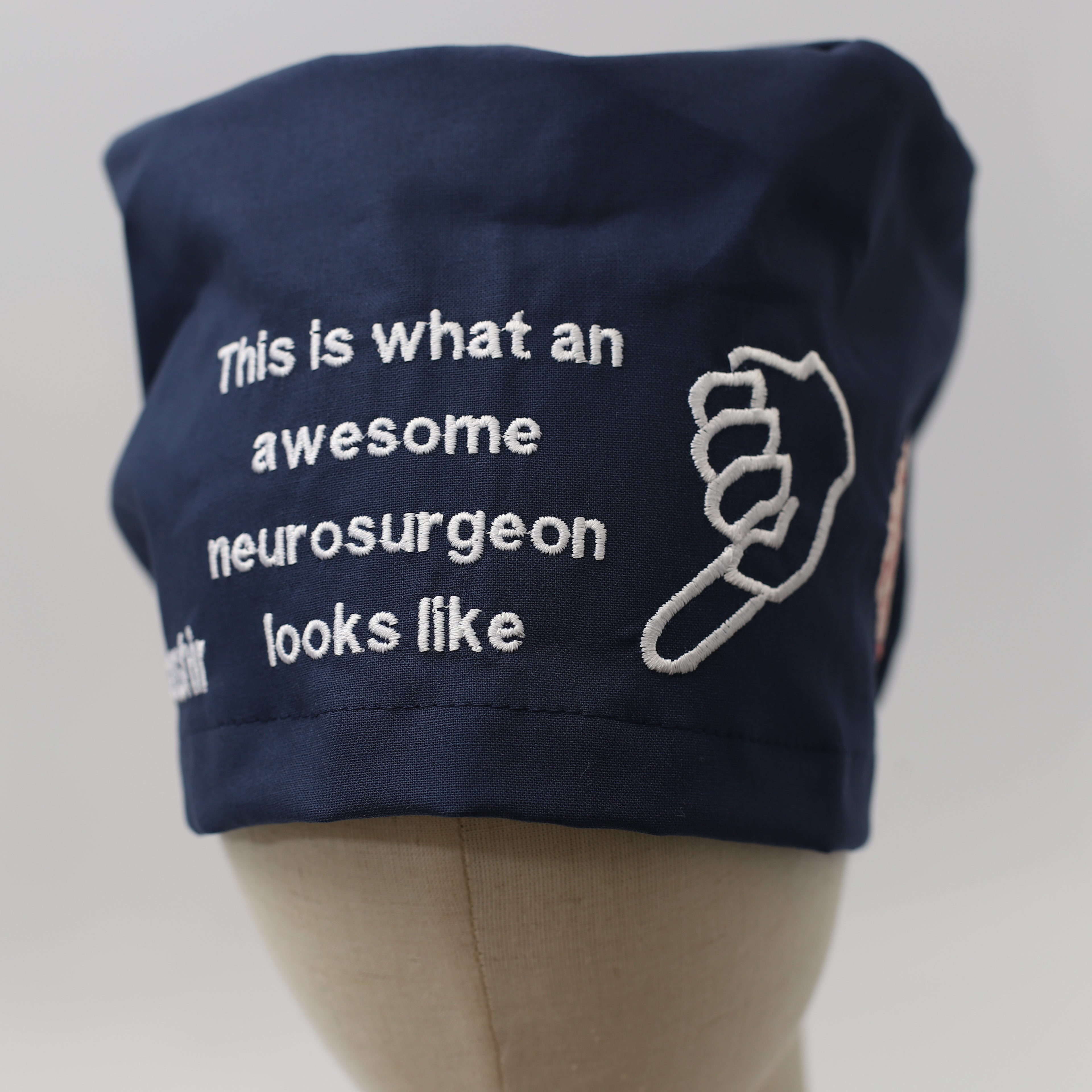 Navy blue Medicus Scrub Cap with white embroidery reading "This is what an awesome neurosurgeon looks like" alongside a pointing hand design.