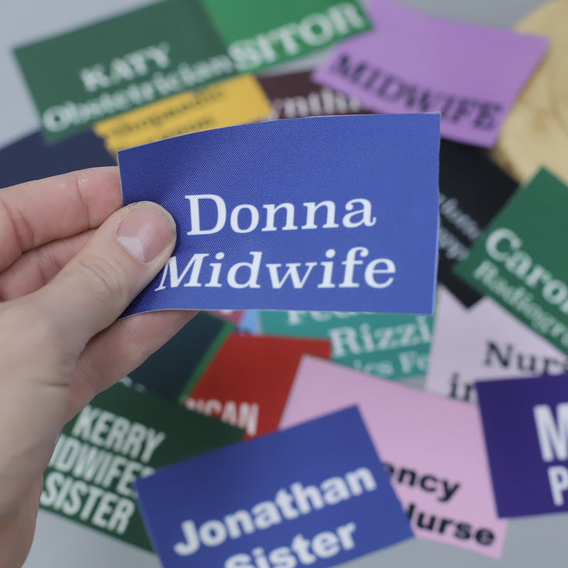 A hand holding a blue card with Donna Midwife printed on it. Various colorful name badges with titles like Sister and Nurse are scattered in the background.
