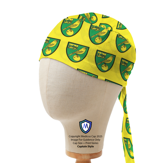 A mannequin head displays a Medicus Scrub Caps Norwich City FC Scrub Cap, featuring a yellow background with green shield motifs and long fabric ties.