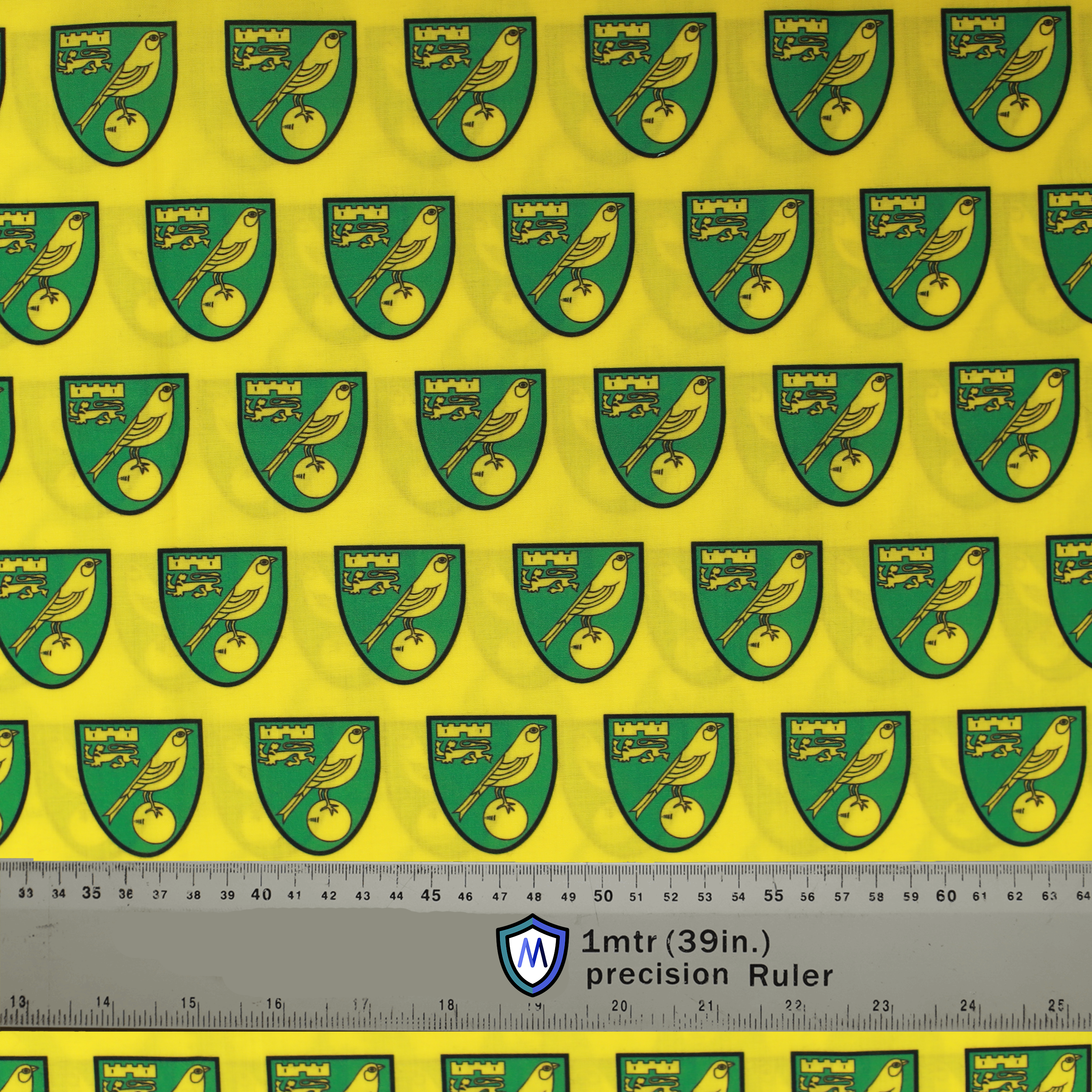 Fabric sample for Medicus Scrub Caps featuring Norwich City FC crests in green and gold on a yellow background, with a 1-meter ruler for scale.
