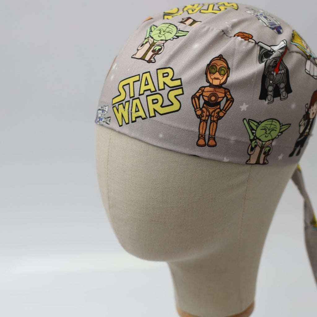 Custom grey scrub cap by Medicus Caps featuring cartoon-style galaxy heroes and robots on a grey background; side view on mannequin with ties.