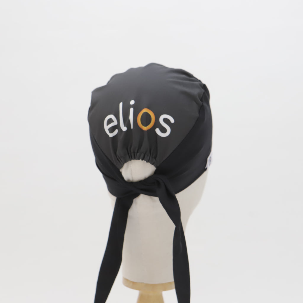 A black custom Medicus Scrub Caps Mullet scrub cap, featuring "elios" branding in white and orange, tied at the back, displayed on a mannequin.