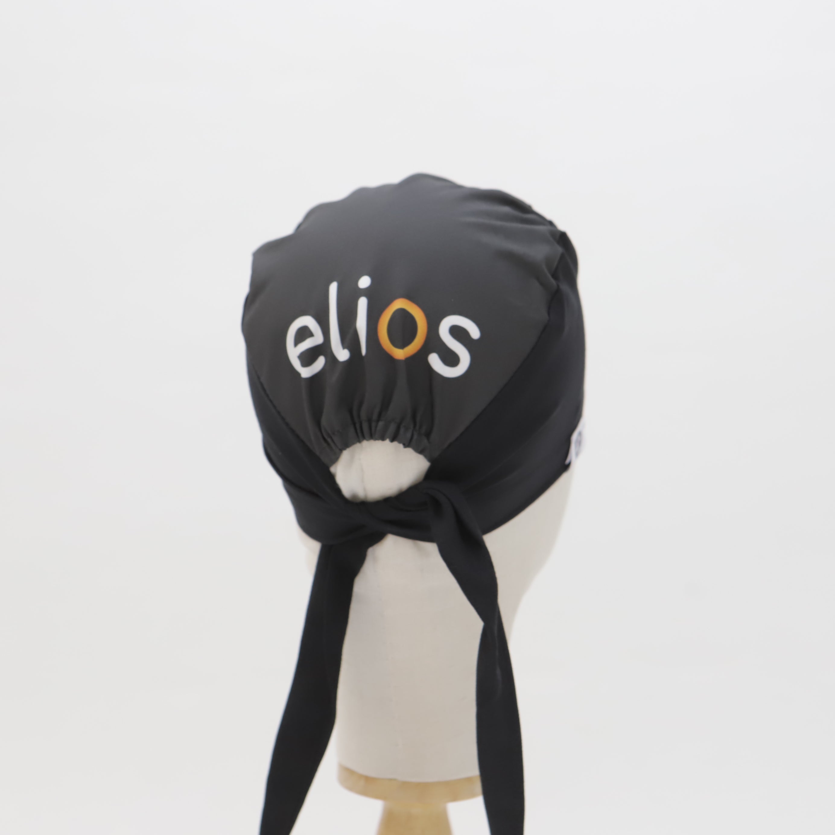 A black custom Medicus Scrub Caps Mullet scrub cap, featuring "elios" branding in white and orange, tied at the back, displayed on a mannequin.