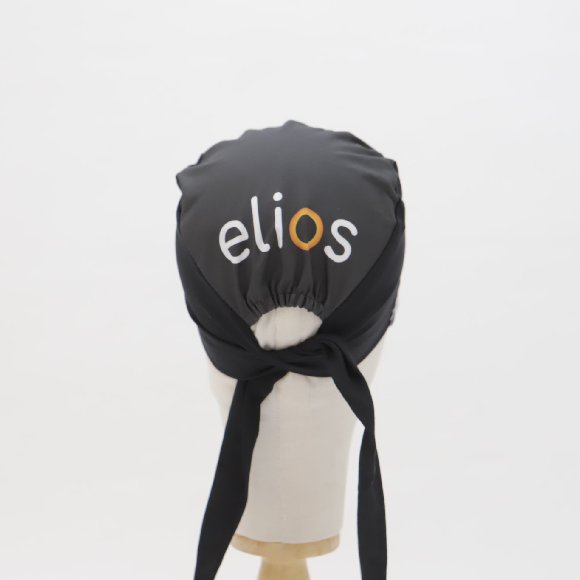 A black Specific Custom Printed Placement Scrub Cap by Medicus Scrub Caps, featuring "elios" in white and orange, displayed on a mannequin against a white background.