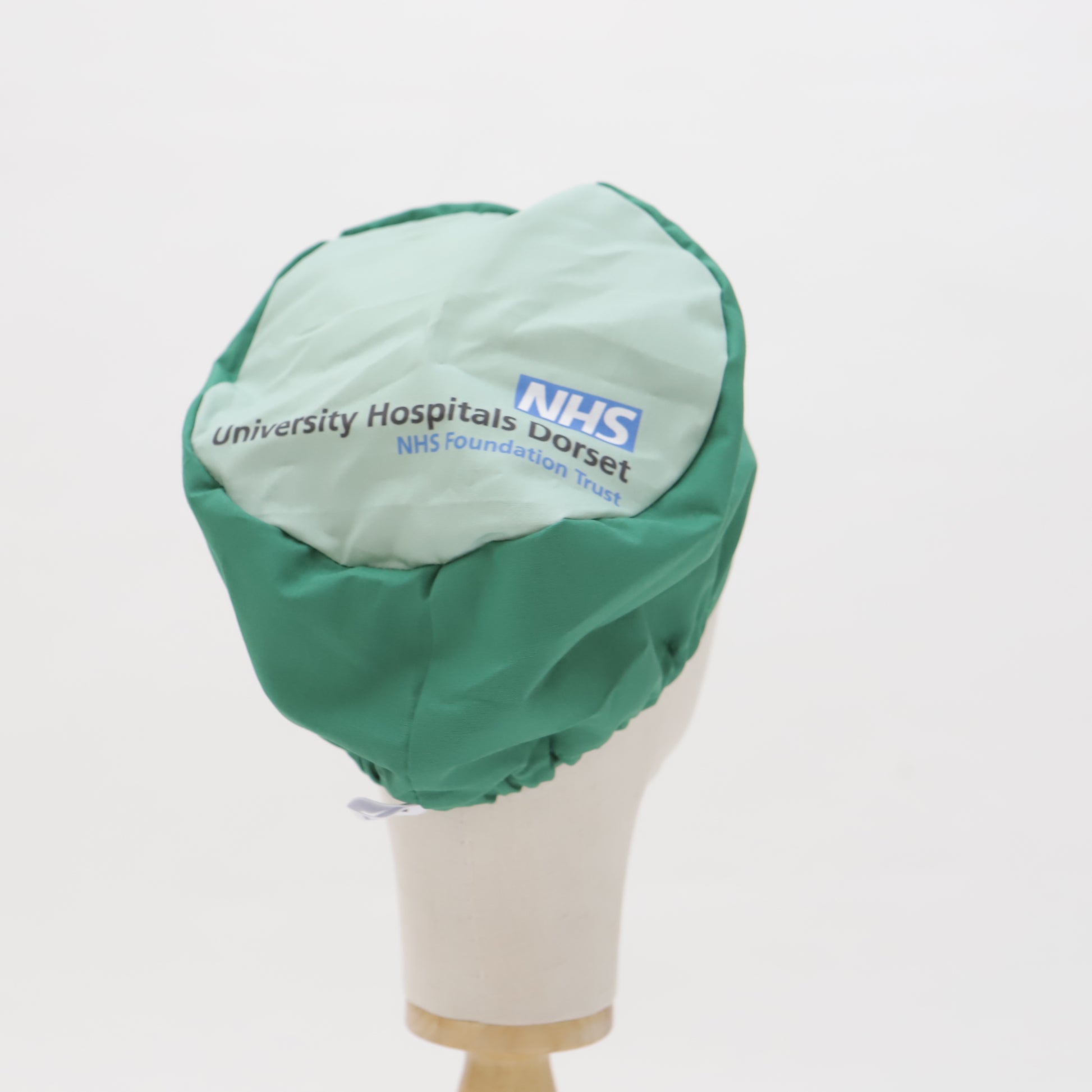 A mannequin displays a green and light blue University Hospitals Dorset NHS scrub cap from Medicus Scrub Caps, a Specific Custom Printed Placement Scrub Cap.