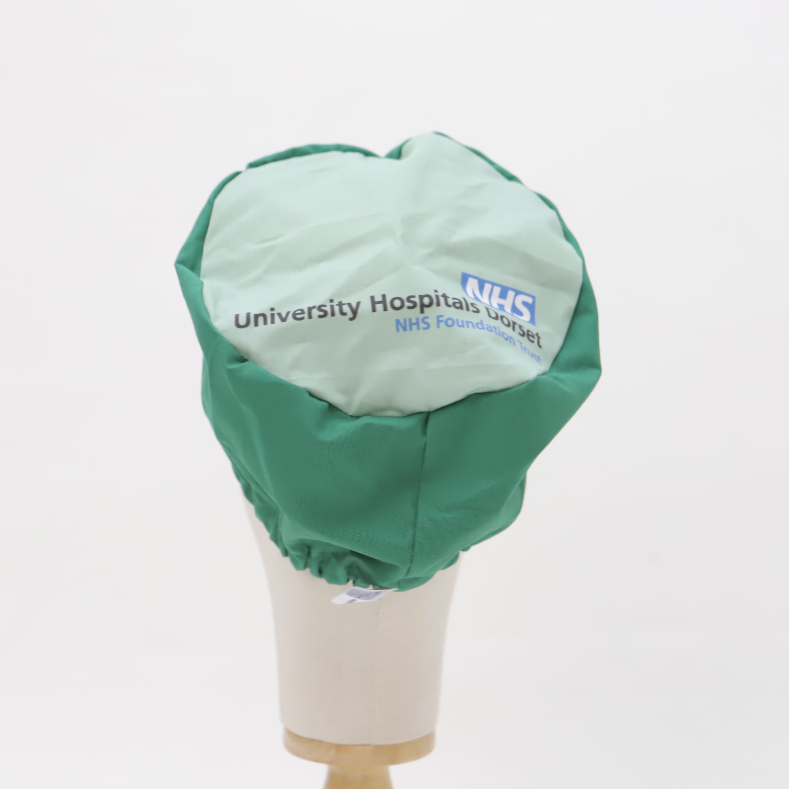 A mannequin head displays a green and light blue custom Mullet Version scrub cap by Medicus Scrub Caps, featuring the University Hospitals Dorset NHS Foundation Trust logo.