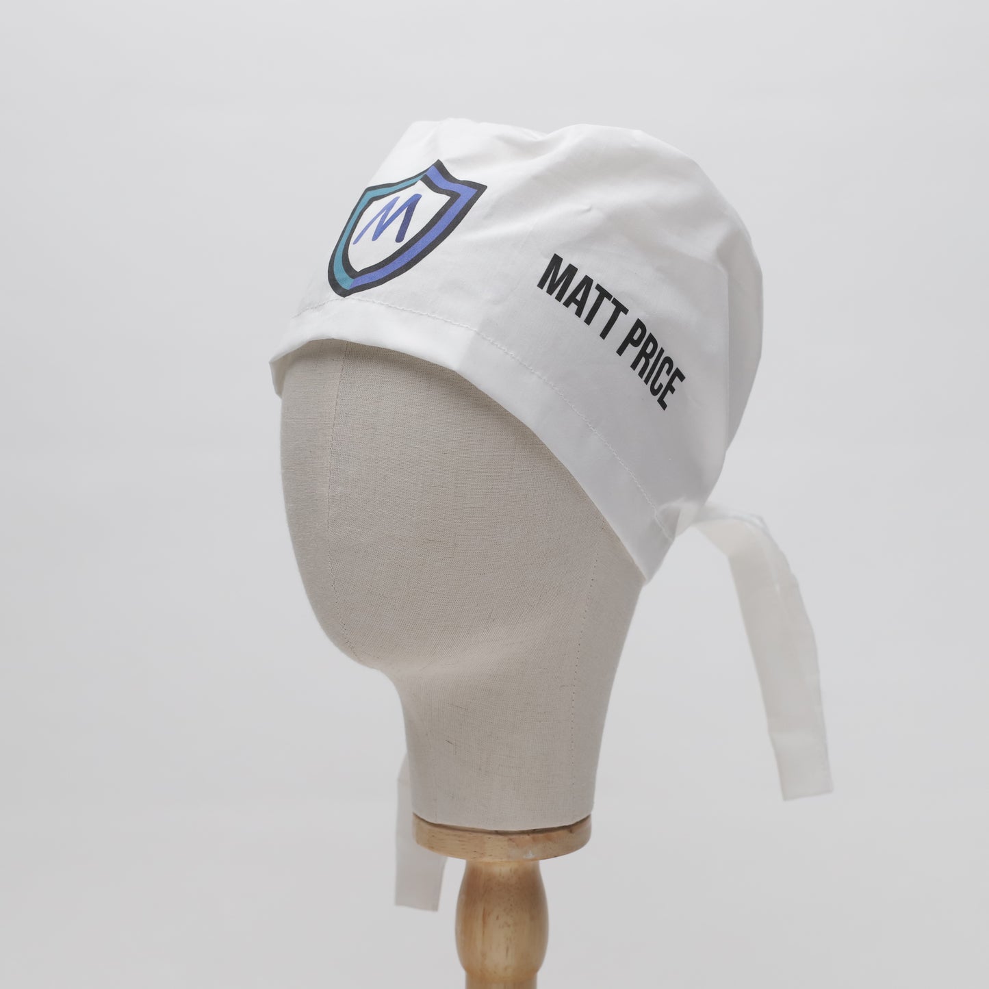 A white mannequin head displays a Medicus Scrub Caps Specific Custom Printed Placement Scrub Cap with a logo and name, featuring long ties for a secure fit.