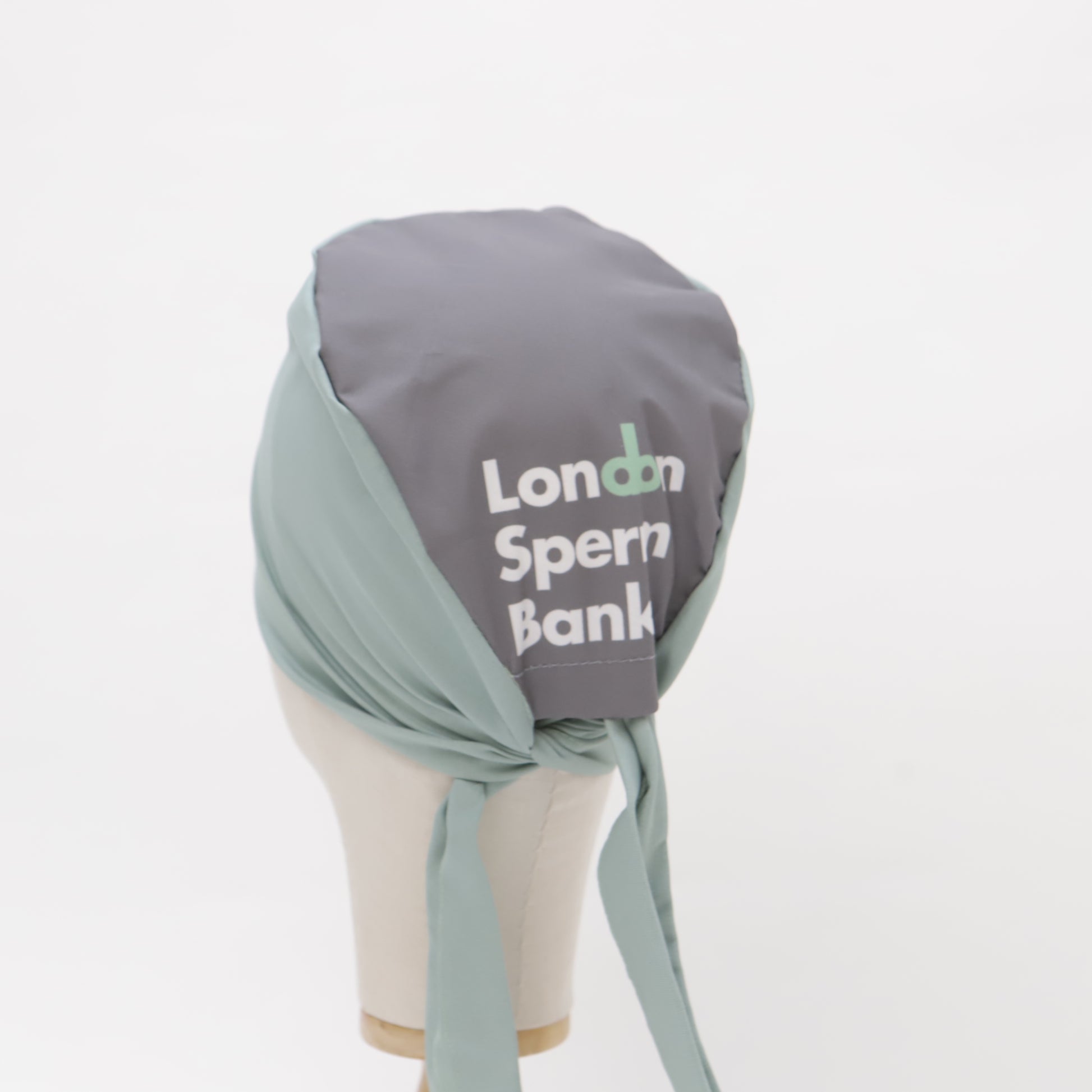 A mannequin head displays a Medicus Scrub Caps scrub cap in light green and gray, custom printed with "London Sperm Bank," neatly tied at the back.