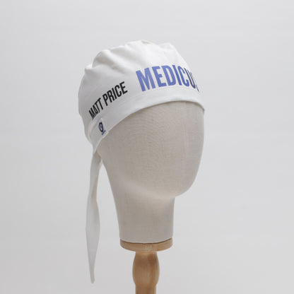 A white mannequin head displays a Medicus Scrub Caps Specific Custom Printed Placement Scrub Cap, featuring MATT PRICE and MEDICUS text, tied at the back on a wooden stand.