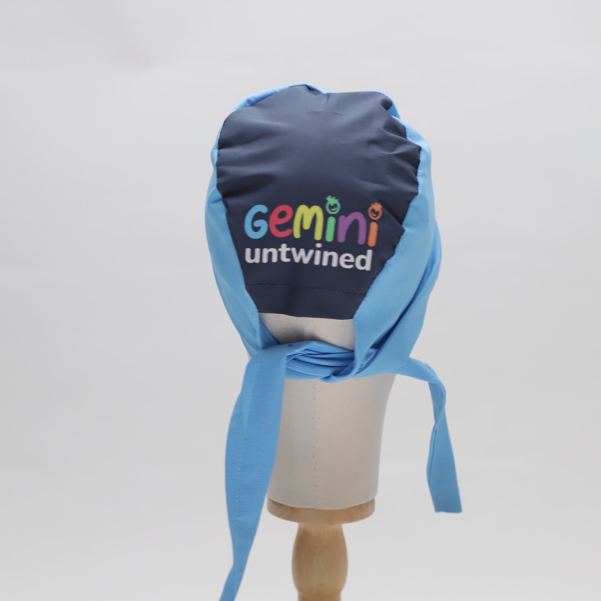 A Medicus Scrub Caps mannequin displays a Specific Custom Printed Placement Scrub Cap in blue and navy with Gemini Untwined text, featuring a stylish tied back.