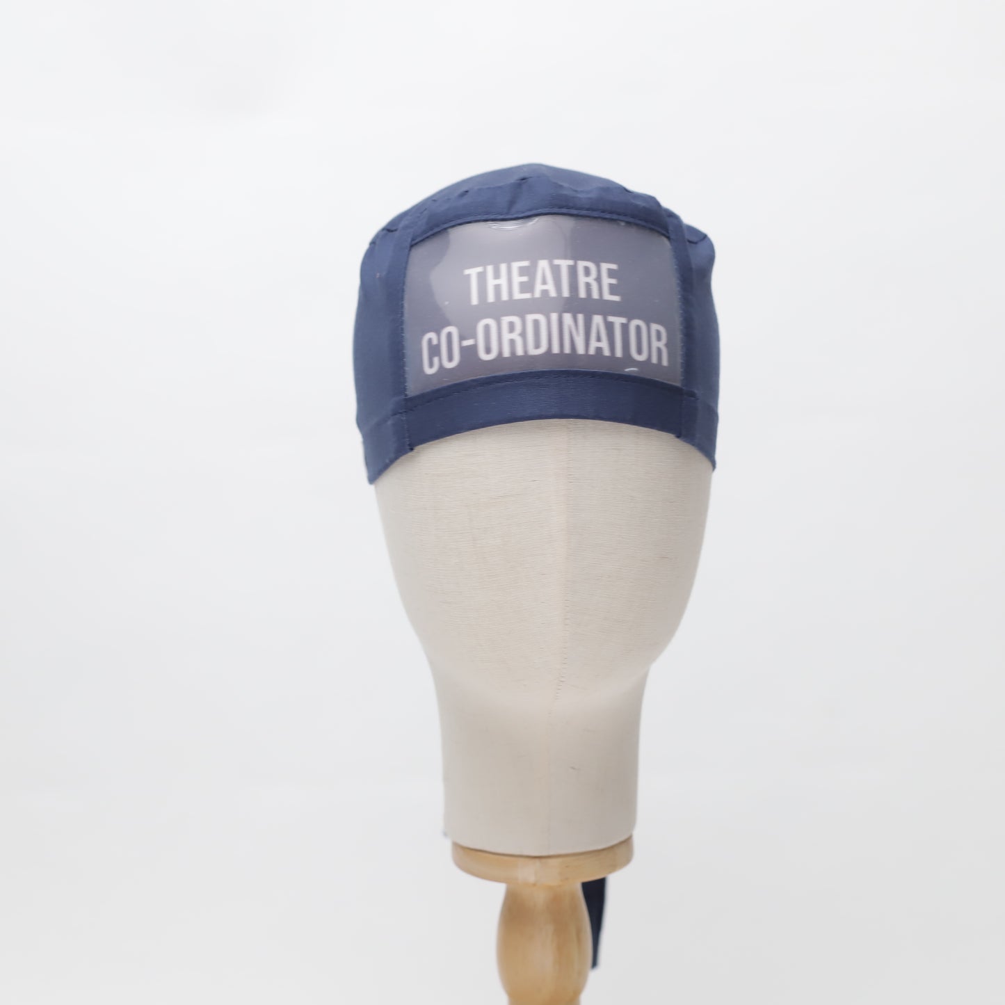 A mannequin head displays a navy Medicus ClearName Scrub Cap with a transparent panel reading "THEATRE CO-ORDINATOR." This reusable cap comes in 22 colors.