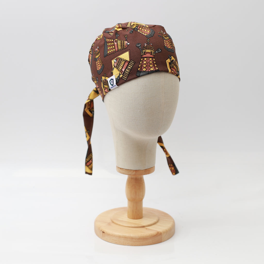 A mannequin head displays a Medicus Scrub Cap in Dalek Brown with a Dr Who-themed pattern, featuring adjustable ties, on a wooden stand against a white background.