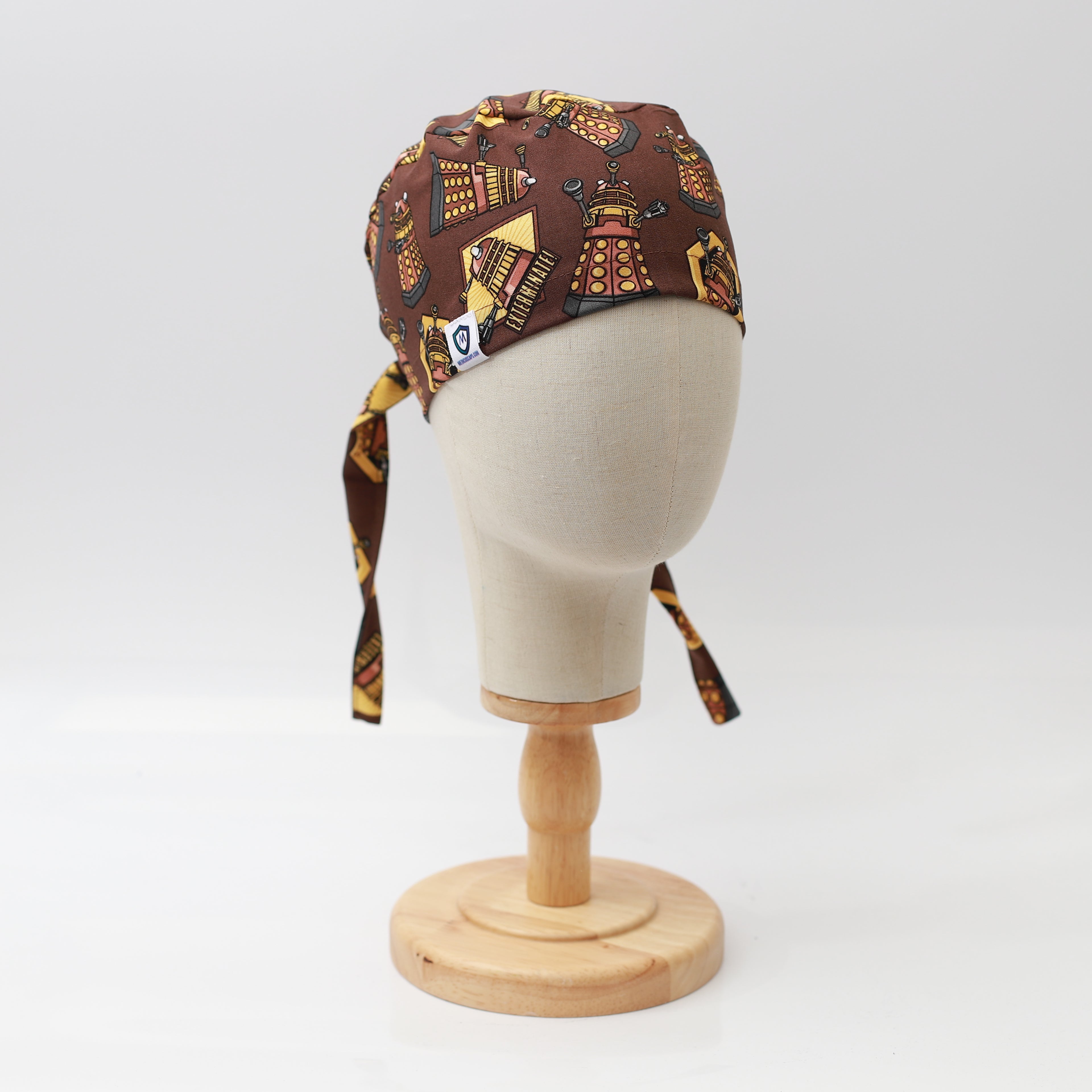 A mannequin head displays a Medicus Scrub Cap in Dalek Brown with a Dr Who-themed pattern, featuring adjustable ties, on a wooden stand against a white background.
