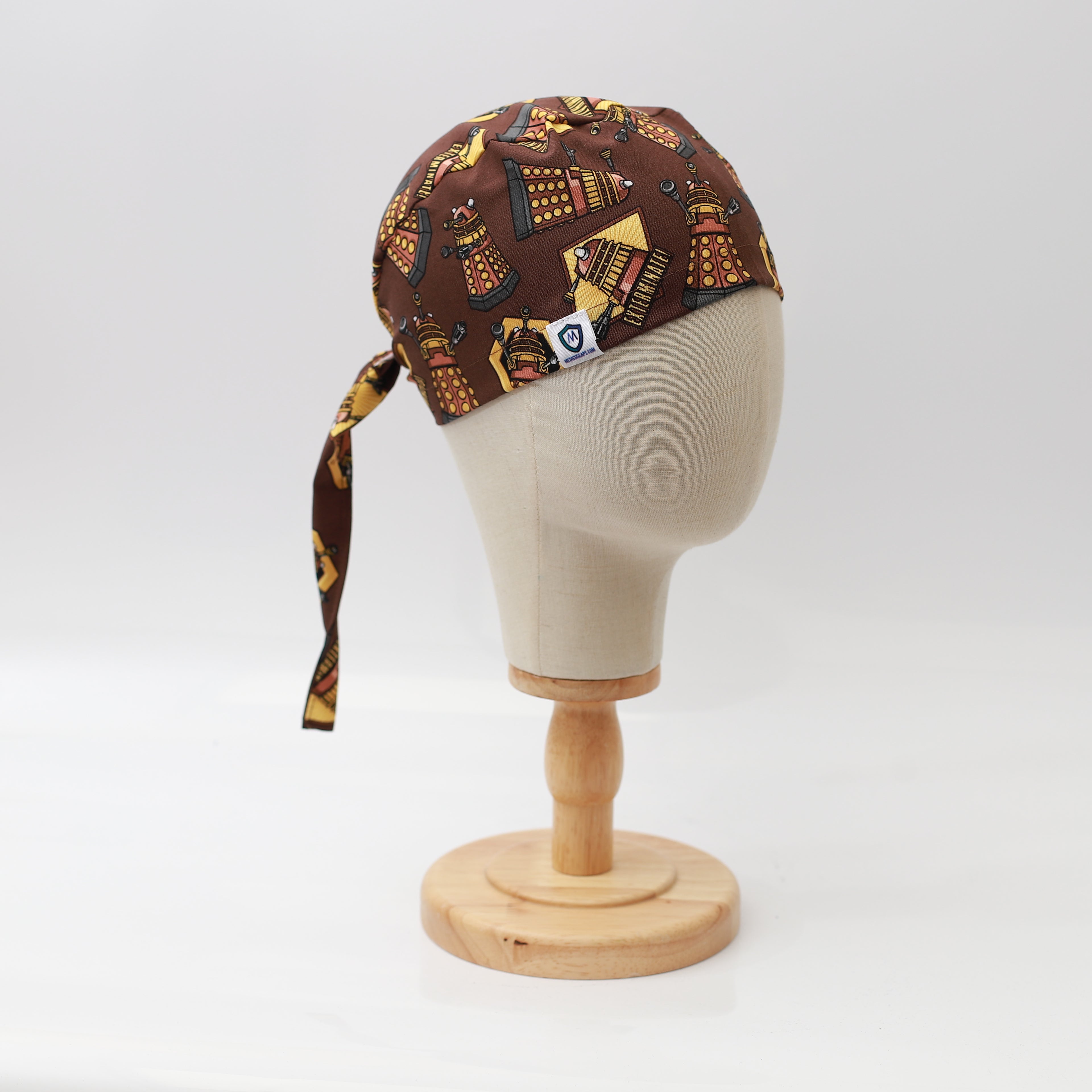 A mannequin displays the Medicus Scrub Caps Dr Who Dalek Brown Scrub Cap, featuring Dalek-themed patterns on brown fabric, tied at the back on a wooden stand.