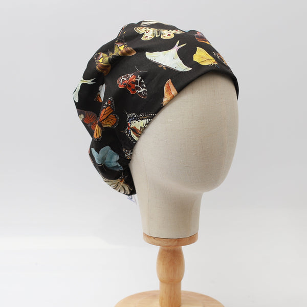 A mannequin head displays the Butterflies Scrub Cap by Medicus Scrub Caps, featuring colorful butterfly patterns on black fabric, elegantly set on a wooden stand.