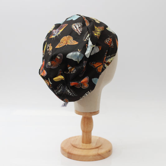 A mannequin head displays the Butterflies Scrub Cap by Medicus Scrub Caps, featuring colorful butterfly patterns on black fabric, set on a wooden stand.