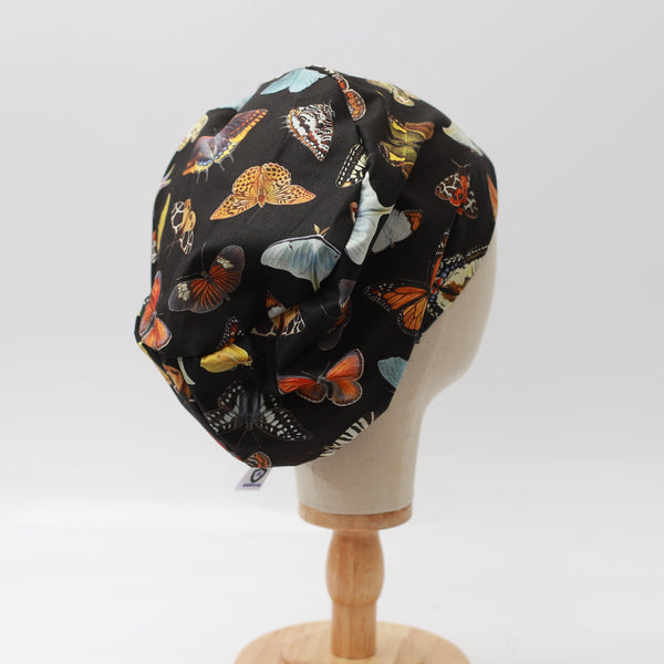 A mannequin head displays the Butterflies Scrub Cap by Medicus Scrub Caps, featuring colorful butterfly patterns on black fabric, set on a wooden stand.