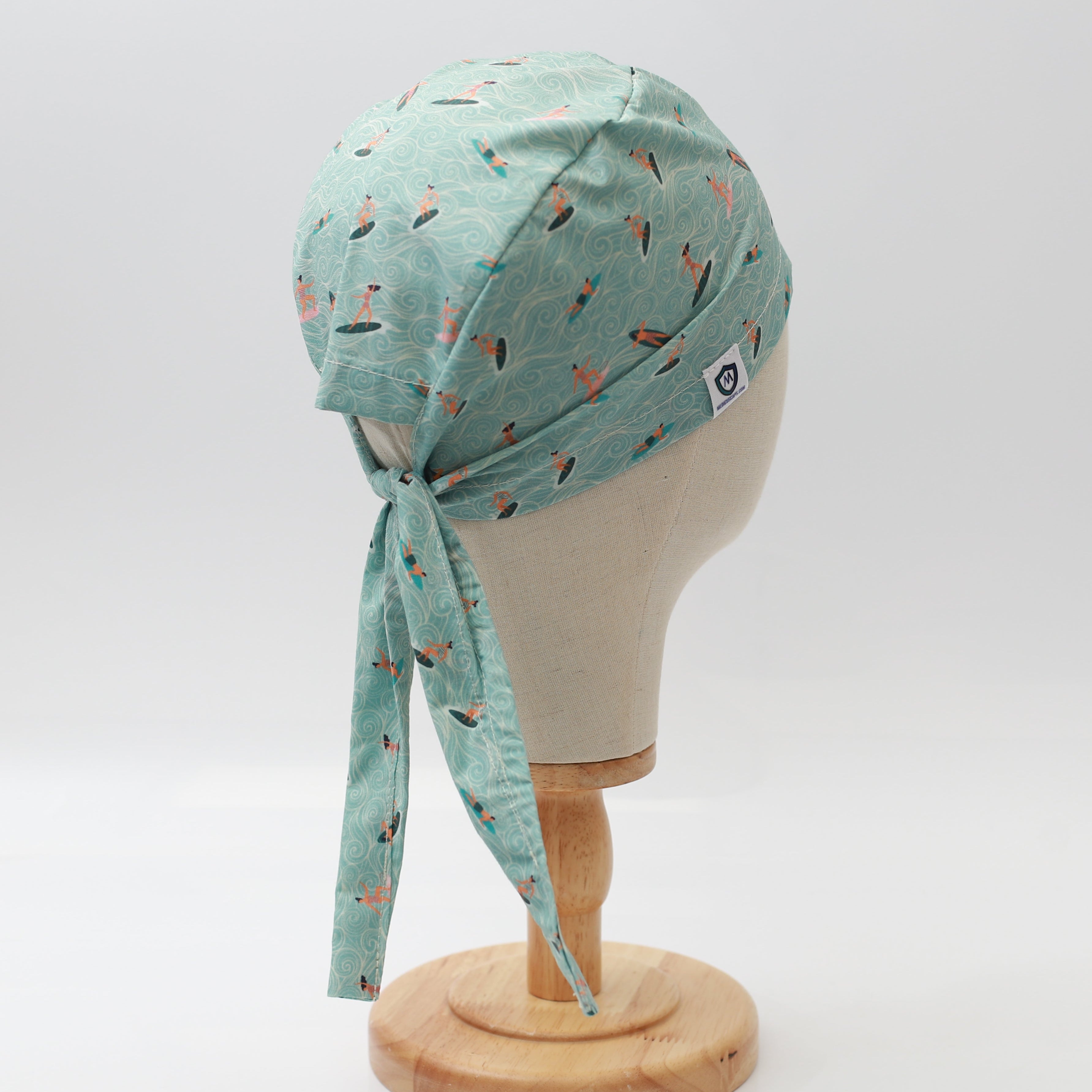 A mannequin displays a Medicus Scrub Caps headscarf with an anime-inspired surfer waves pattern. Light blue fabric, tied at the back, on a wooden stand.