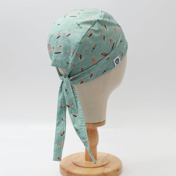 A mannequin displays a Medicus Scrub Caps headscarf with an anime-inspired surfer waves pattern. Light blue fabric, tied at the back, on a wooden stand.