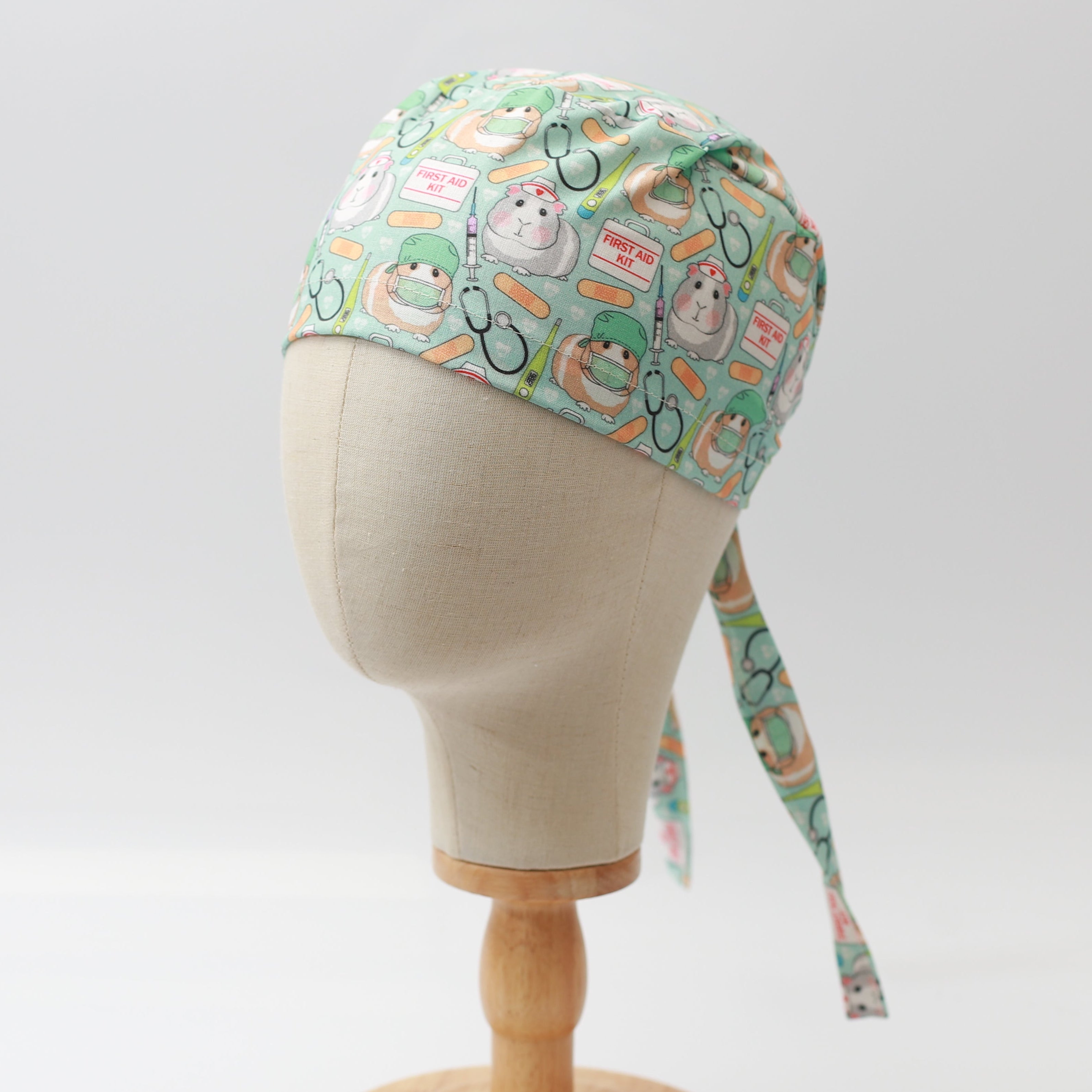 Medicus Scrub Cap with a cute guinea pig doctor and first aid pattern on a light green background, displayed on a mannequin head.