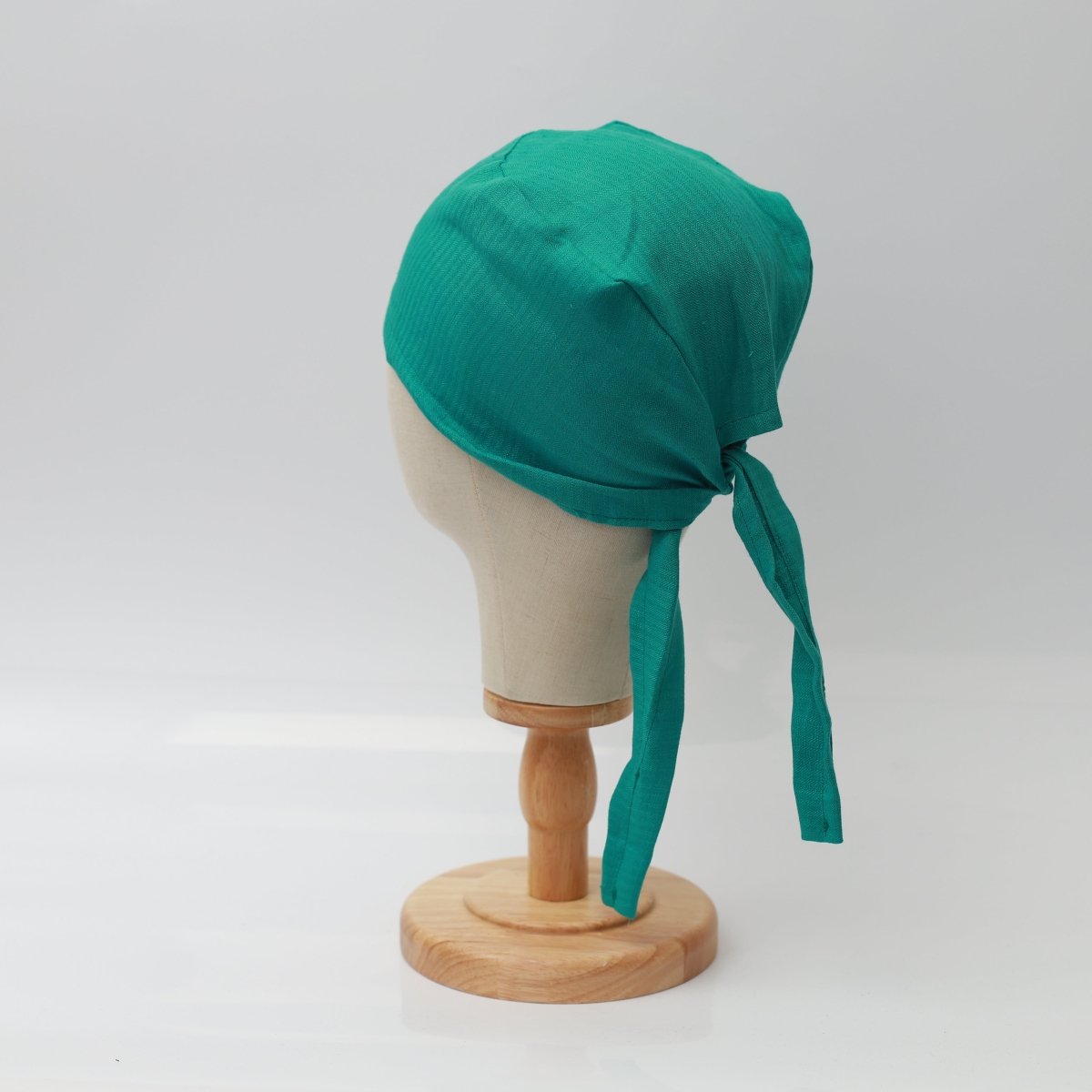 Displayed on a wooden stand, a mannequin head wears the Medicus Scrub Caps 100% Hemp Fabric Reusable Scrub Cap in teal, tied at the back.