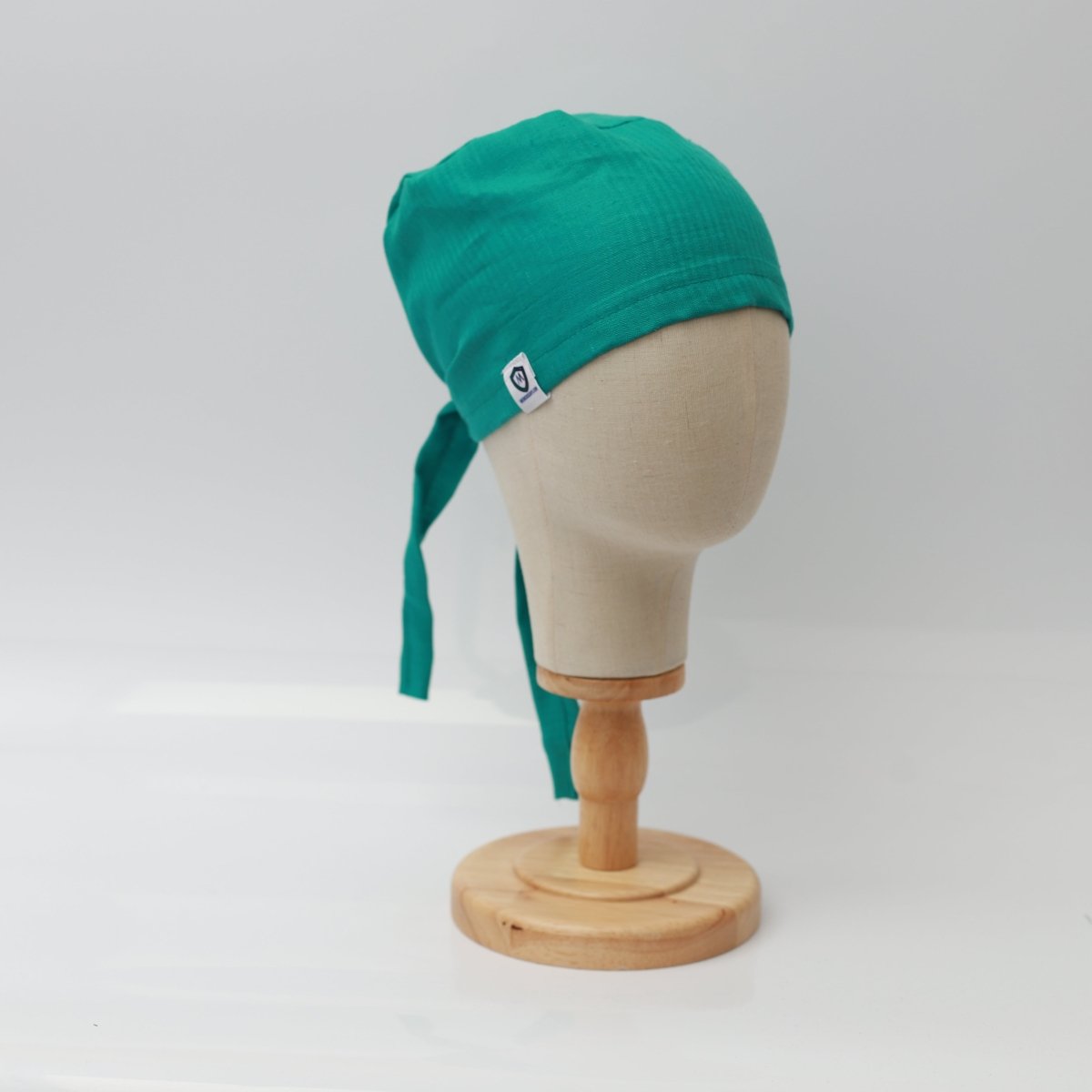 A mannequin head displays the Medicus Scrub Caps teal green 100% hemp fabric reusable scrub cap with long ties and a white tag, on a wooden stand.