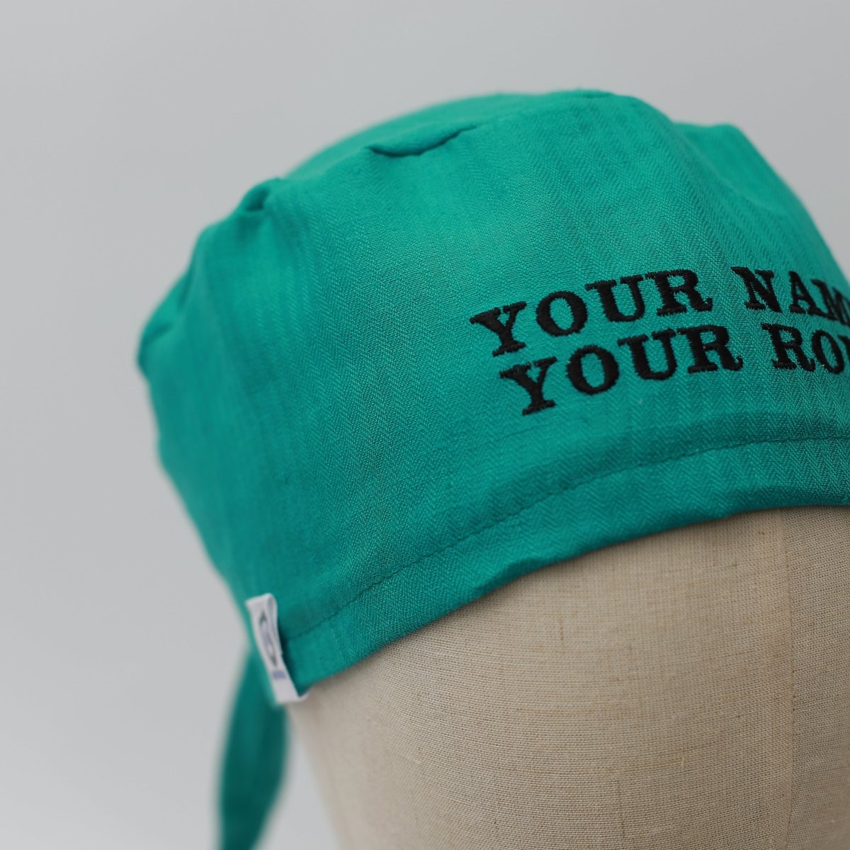 The Medicus Scrub Caps teal green surgical cap, made from 100% hemp fabric, features black embroidered text and a reusable design, modeled on a mannequin.