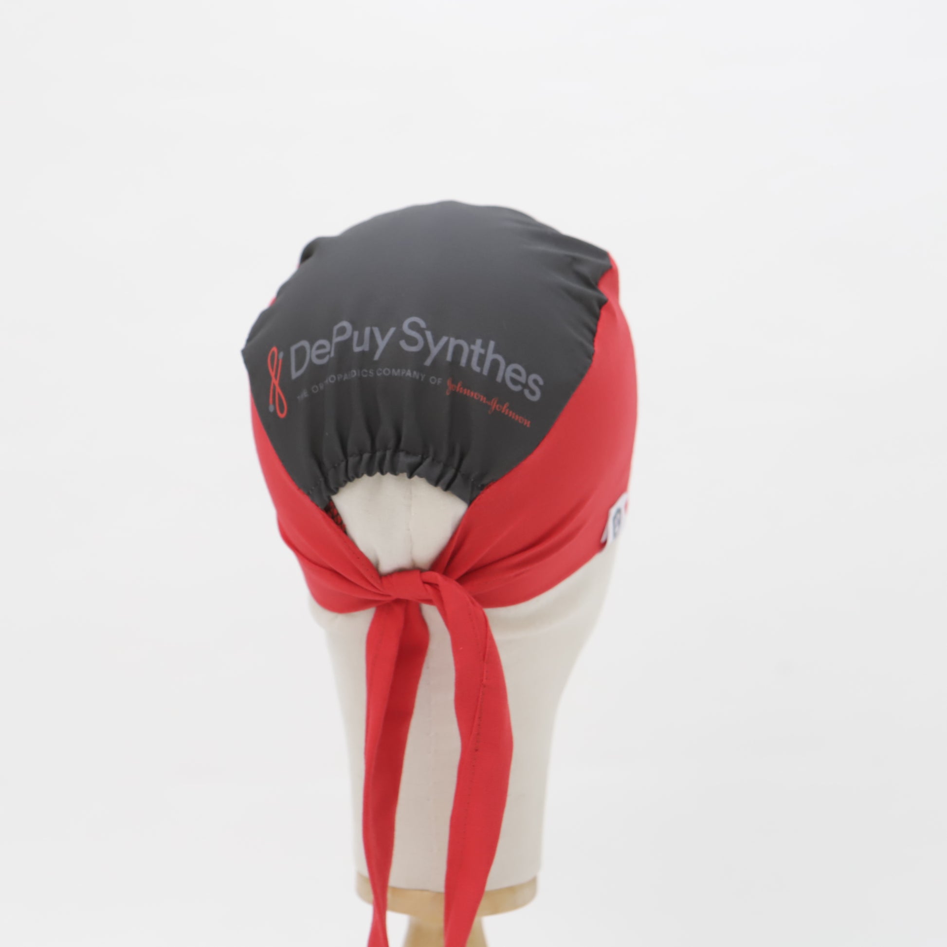 A mannequin head displays a black and red DePuy Synthes scrub cap by Medicus Scrub Caps, featuring a back-tied bow and custom-printed design.