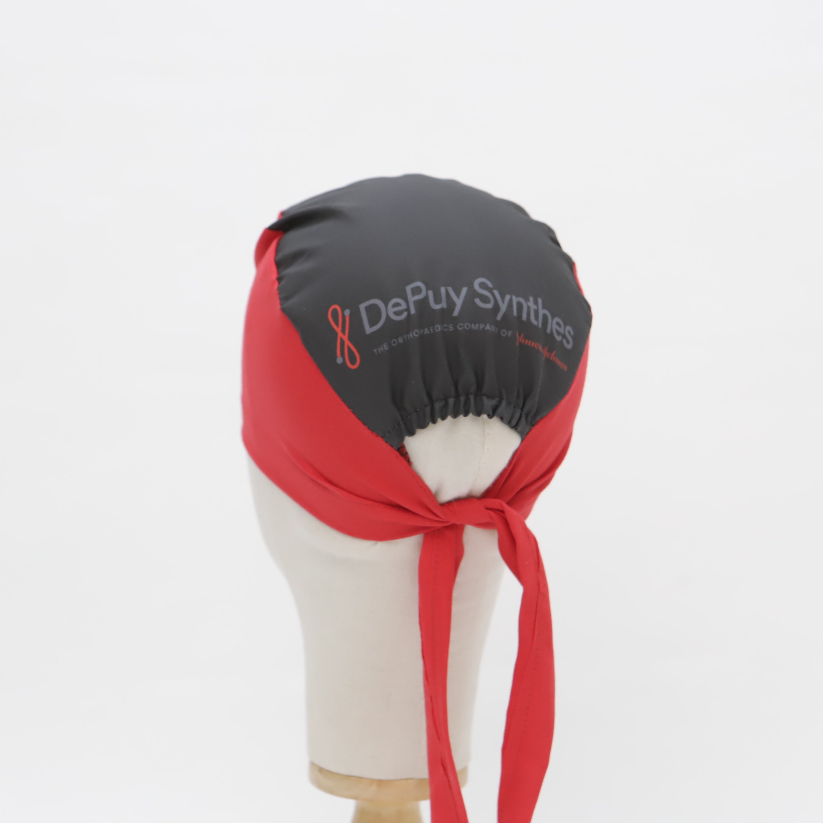 A mannequin head displays a black Medicus Scrub Cap with the DePuy Synthes logo, featuring a red tie-back "mullet" design against a plain white background.