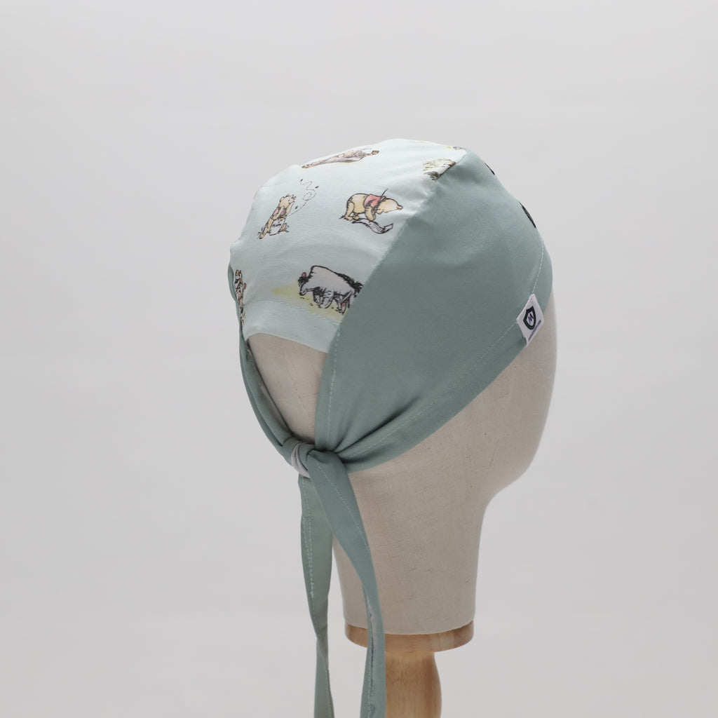 A mannequin displays The Mullet scrub cap by Medicus Scrub Caps, featuring light green fabric with illustrated animals and fabric ties at the back.