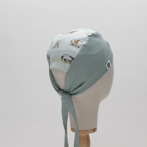 A mannequin displays The Mullet scrub cap by Medicus Scrub Caps, featuring light green fabric with illustrated animals and fabric ties at the back.
