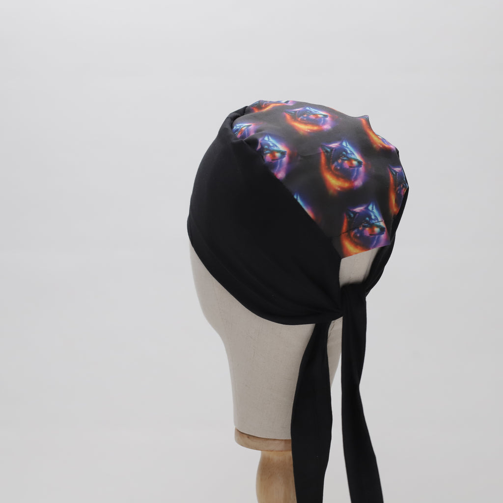 A mannequin head displays The Mullet Scrub Cap by Medicus Scrub Caps, featuring a black base with a vibrant abstract pattern and long ties at the back.