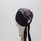 A mannequin head displays The Mullet Scrub Cap by Medicus Scrub Caps, featuring a black base with a vibrant abstract pattern and long ties at the back.