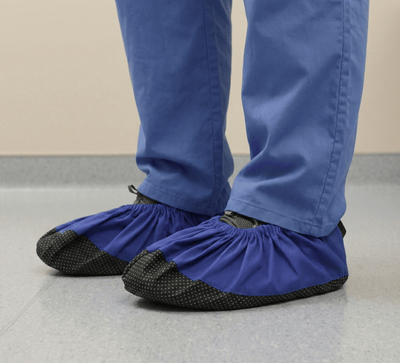 Navy blue reusable shoe covers with black textured soles worn over medical scrubs