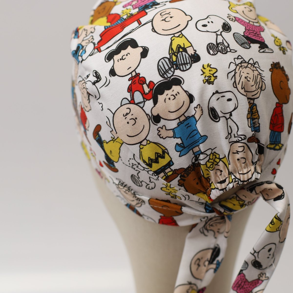 Bouffant style custom scrub cap featuring classic Peanuts cartoon characters like Charlie Brown, Lucy, and Snoopy on a white background with colorful outfits, handmade by Medicus Caps.