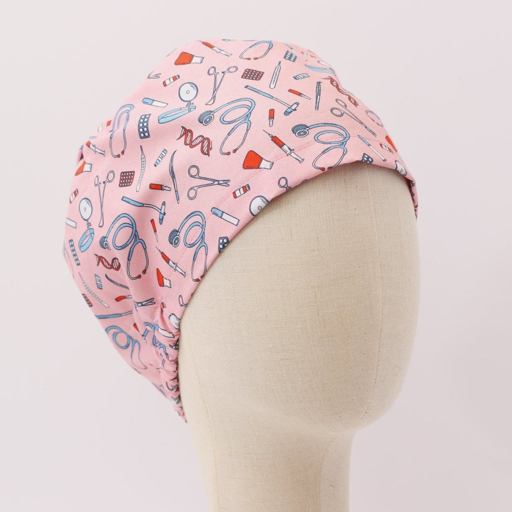 Custom pink scrub cap by Medicus Caps featuring illustrated medical equipment like stethoscopes, syringes, scissors and pills. Bouffant style surgical cap made to order.