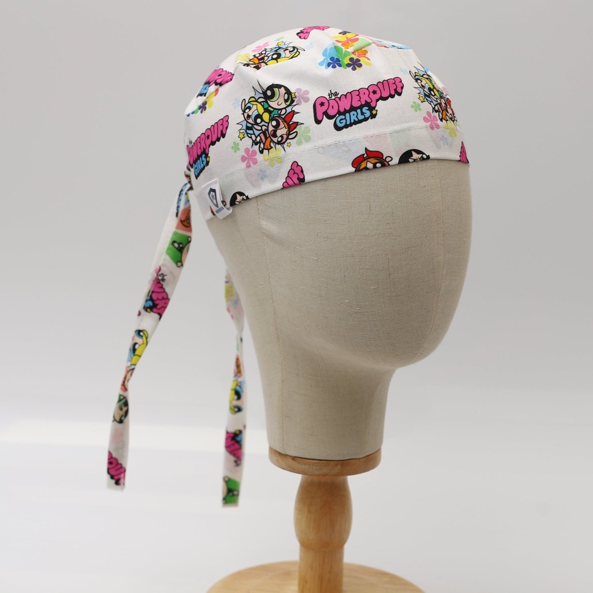 Custom scrub cap with Powerpuff Girls print by Medicus Caps; white fabric with colorful cartoon characters. Side view shows long ties, handmade to order.