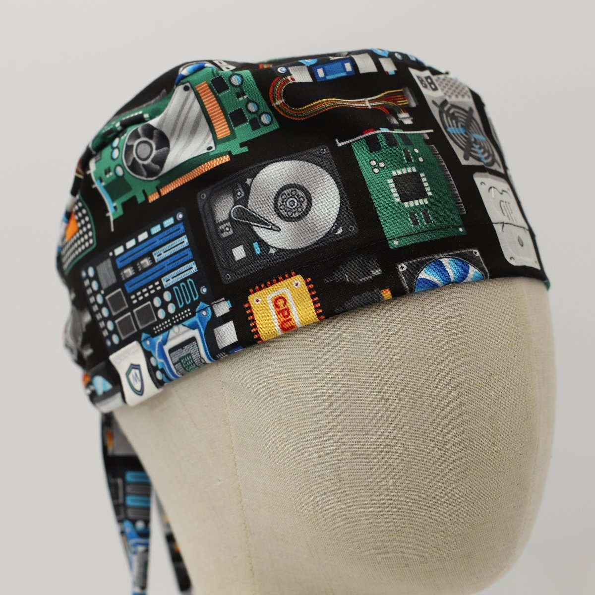 Custom black scrub cap by Medicus Caps featuring a pattern of retro technology and electronics icons, including cassette tapes, circuit boards, and computer hardware; side view on mannequin with long ties.