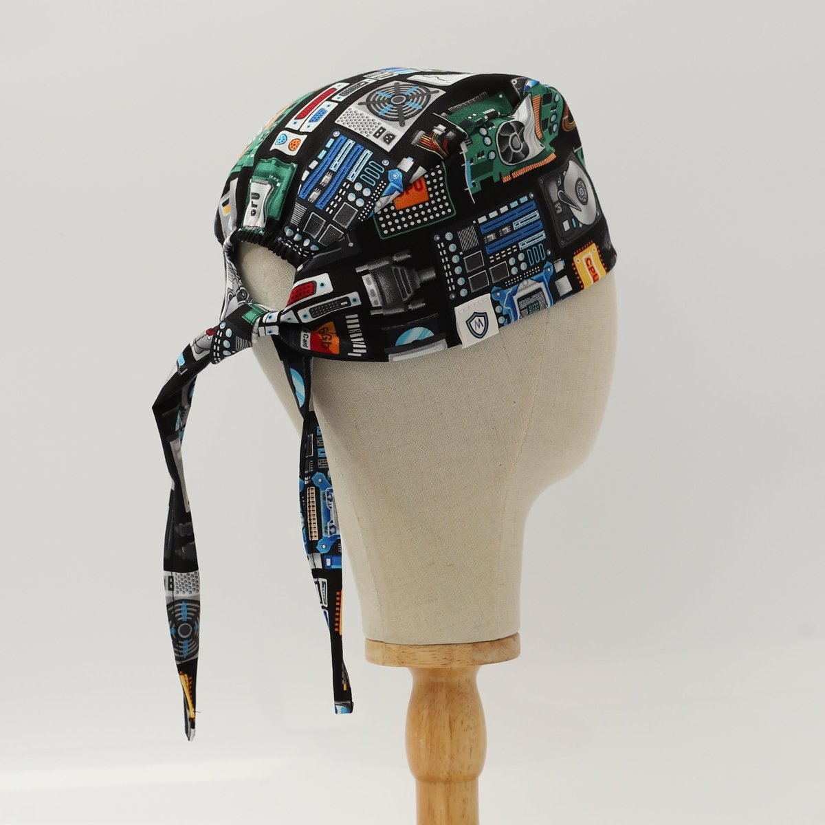 Custom black scrub cap by Medicus Caps featuring retro technology and electronics print including cassette tapes, circuit boards, and computer hardware; back view showing tie straps on mannequin.