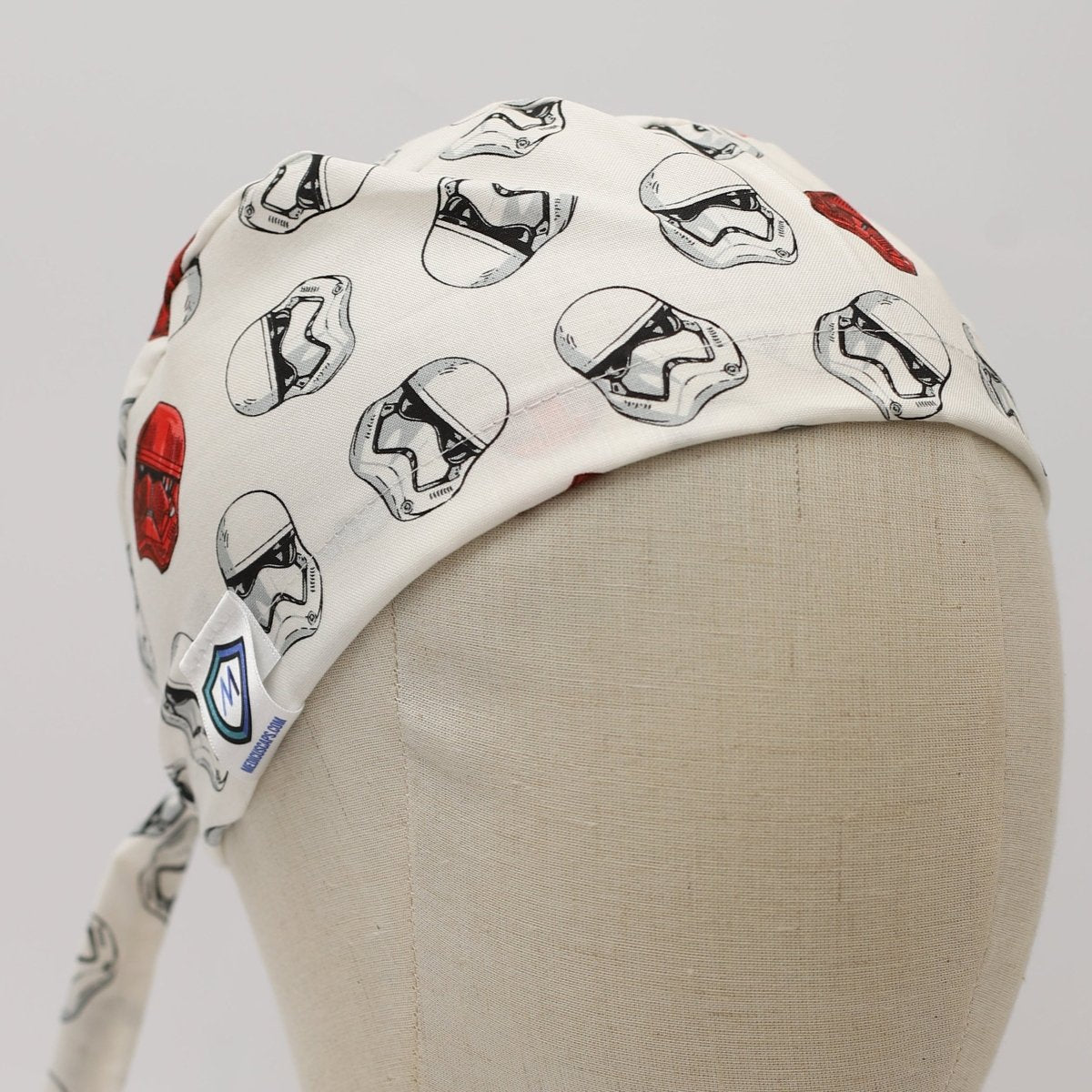 Custom white scrub cap by Medicus Caps featuring repeating black and red sci-fi helmet designs; side view on mannequin with ties.